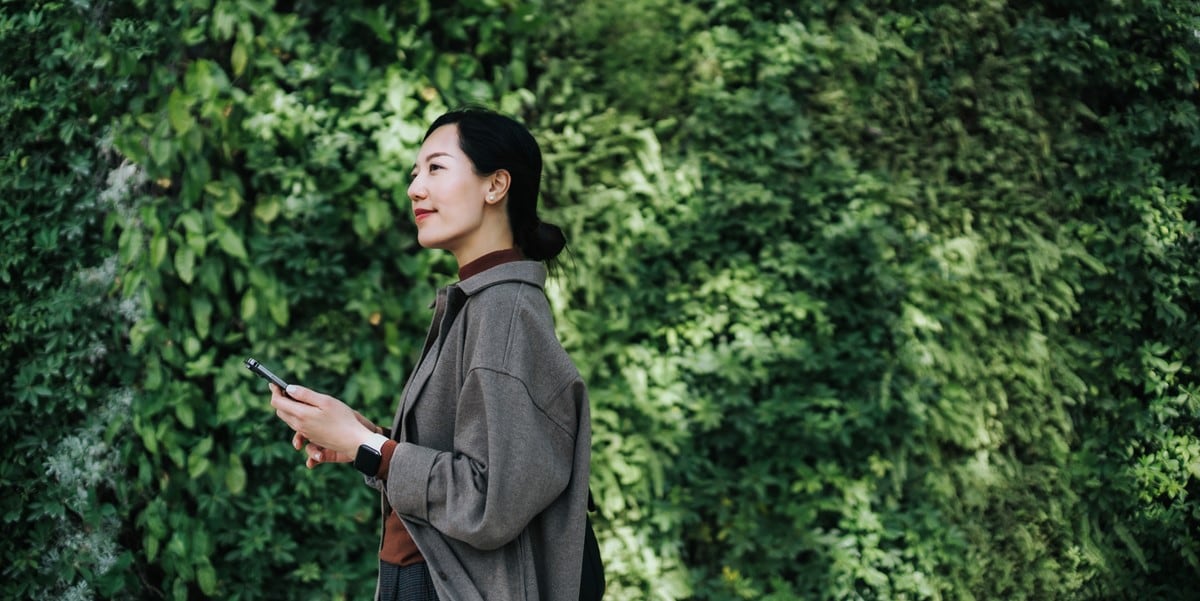 woman standing in front of greenery outside, phone in hand