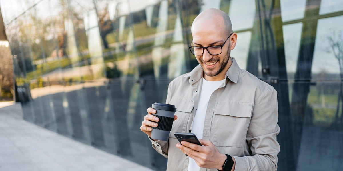 man standing outside building holding coffee cup, looking at cellphone
