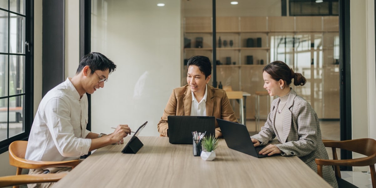 3 professionals seated at a conference table with laptops in front of them