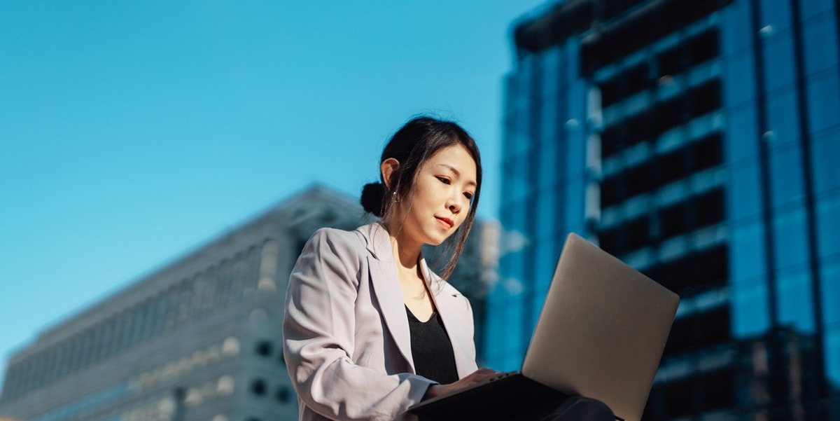 professional woman sitting outside in front of office buildings, using laptop