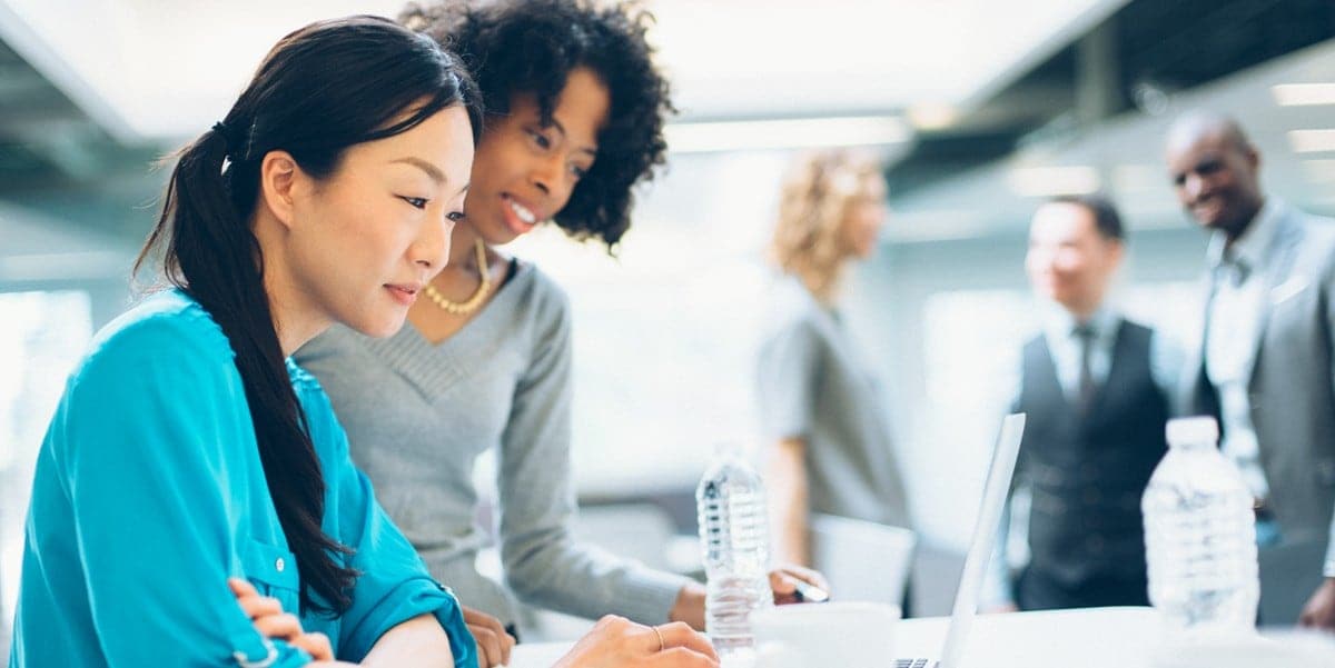 two women sitting at a desk, colleagues in the background