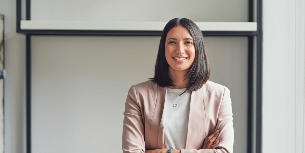 woman in pink blazer smiling, arms crossed, in the office
